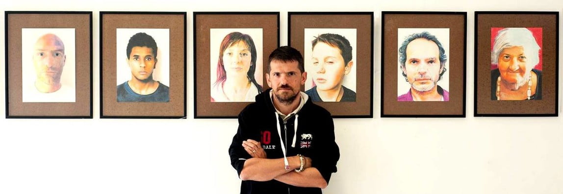 Luciano Blini standing in front of a wall adorned with pictures of people, representing diverse faces and stories.